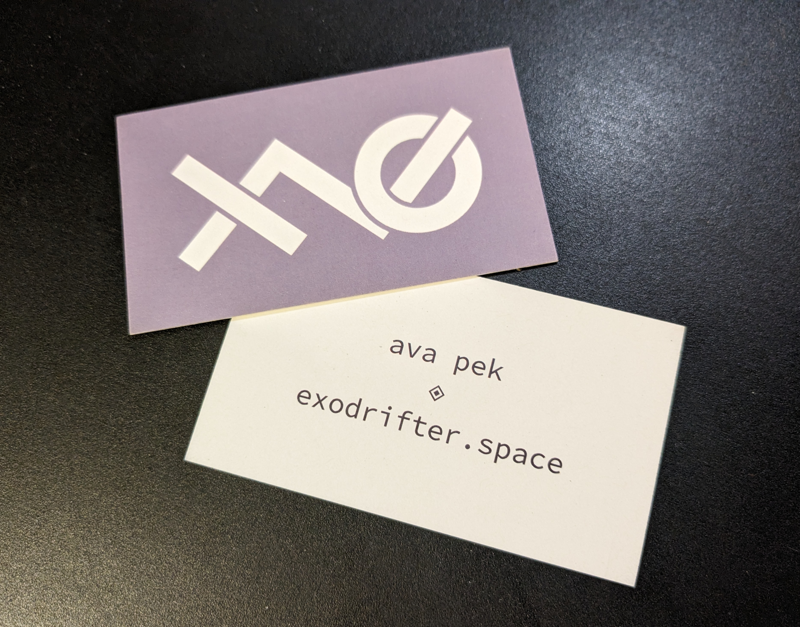 Two business cards on a black surface. The first one shows the front of the business card is a logo, which combines the letters 'x' and 'o' with a tilde in white on a purple-gray background. It's slightly overlapping the second card that is flipped over to show the back of the business card, which reads "ava pek" and "exodrifter.space" on lines separated by two cocentric squares rotated so that their corners point to the edges of the card.