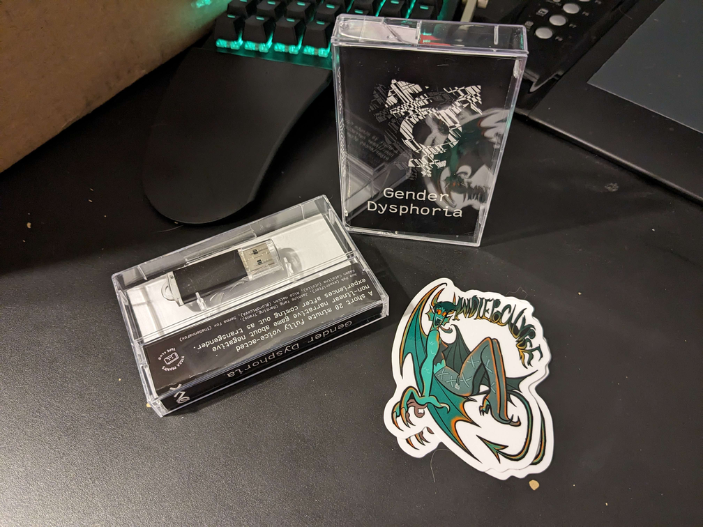 Two cassette cases, one standing upfront showing the cover art for the game Gender Dysphoria and one laying face-down showing the side and back of the cassette case, where a USB is visible instead of a cassette tape. They are both placed on a black desk next to an Indiepocalyspe devil sticker in the foreground and a cardboard box, the left keyboard of a Moonlander, and a cheap Monoprice tablet in the background.