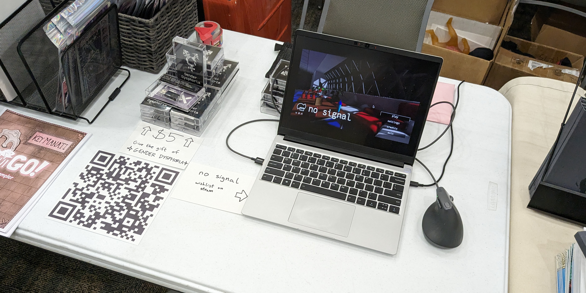 My half of the table at STAPLE! Austin 2024. The table is not covered by a tablecloth. A stack of Gender Dysphoria cassette cases sit to the left, with an index card below it which reads "Give the gift of ⭐ GENDER DYSPHORIA ⭐". A Framework laptop and a wired ergonomic mouse sits to the right, with an index card pointing at it which reads "no signal" and "wishlist on steam"