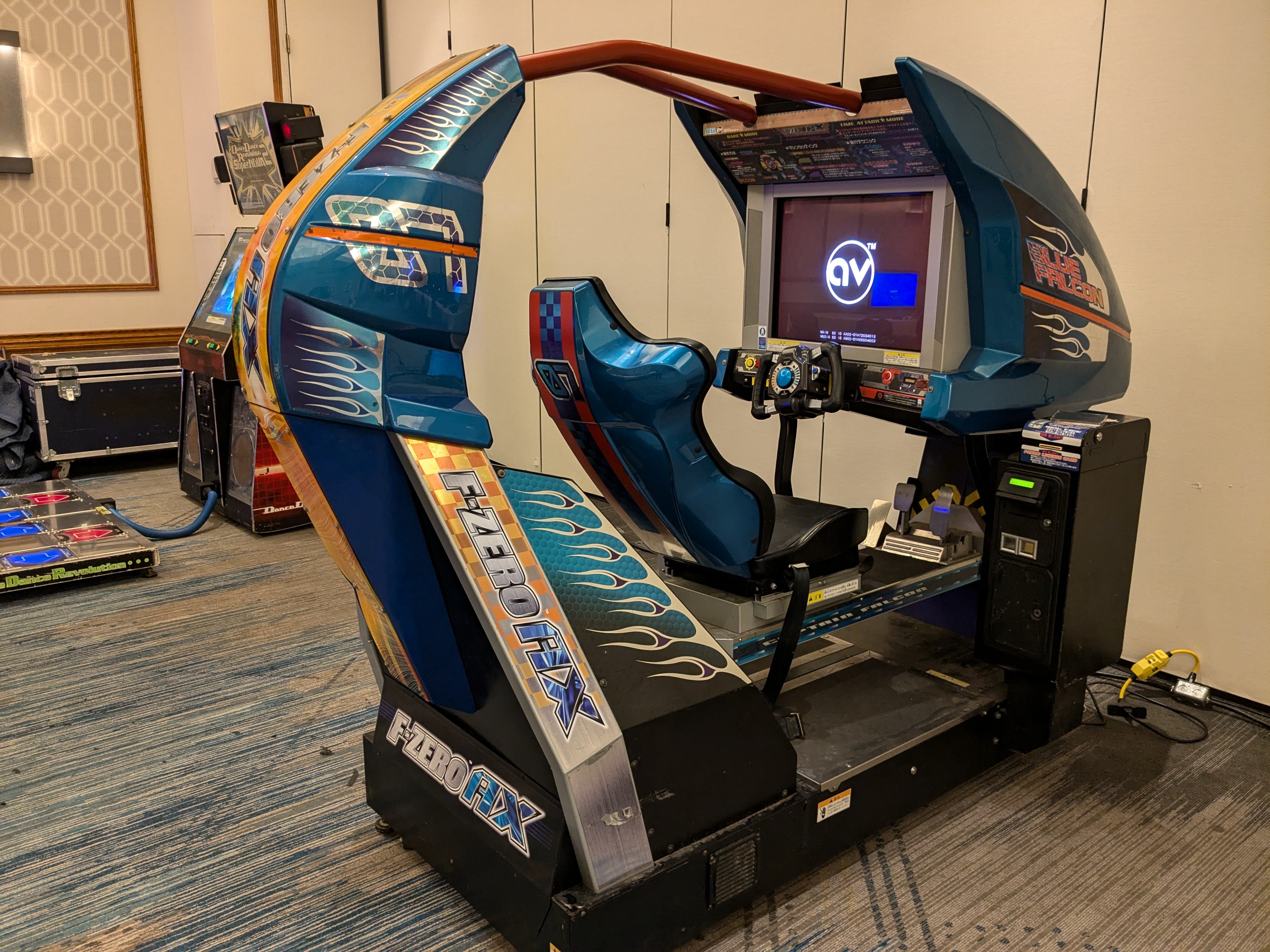 An F-Zero AX arcade cabinet. No one is currently sitting in it. There's a Dance Dance Revolution Supernova 2 arcade cabinet in the background.