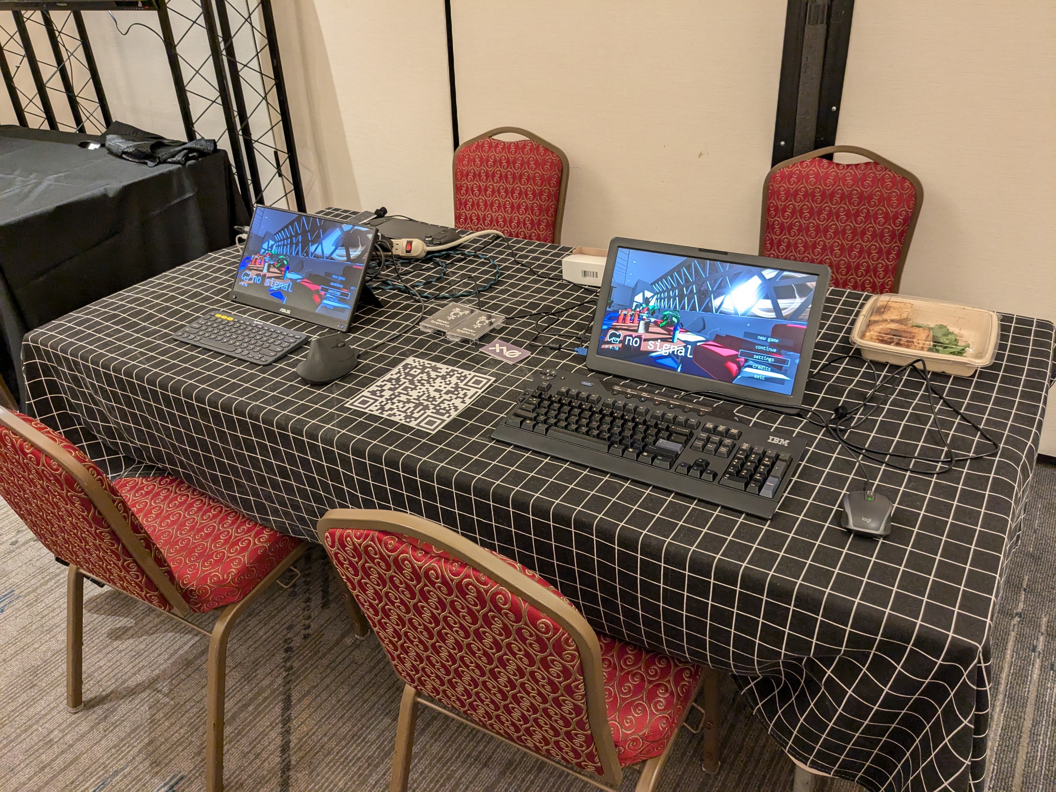 The table setup at Delta H Con. The tablecloth is black, with a white grid pattern. There are two screens with no signal running on them, each with a keyboard and mouse. There are some cassette copies of Gender Dysphoria, a large QR code for the no signal Steam Page, and a boxed mushroom pesto sandwich.