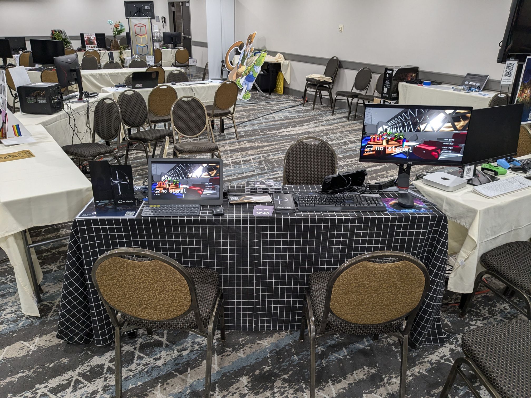 My table at 2D Con 2024. It's covered with a black tablecloth with a white grid pattern. On the table sits two monitors, one of which is portable and the other is not. There are also flyers on the table, one of which is propped up to show the space station from the game.