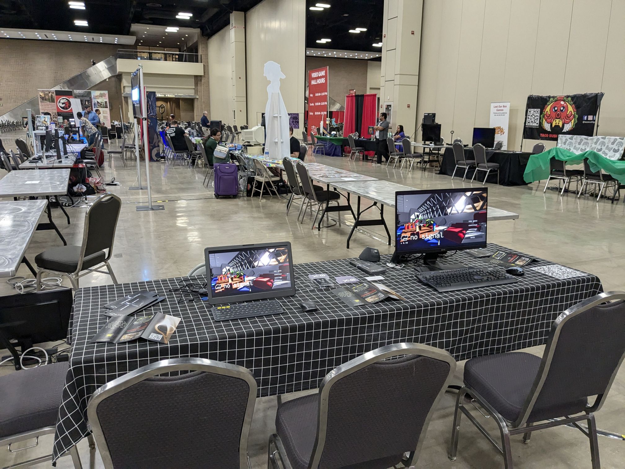 My table at San Japan 2024. It's covered with a black tablecloth with a white grid pattern. On the table sits two monitors, one of which is portable and the other is not. There are also flyers on the table.